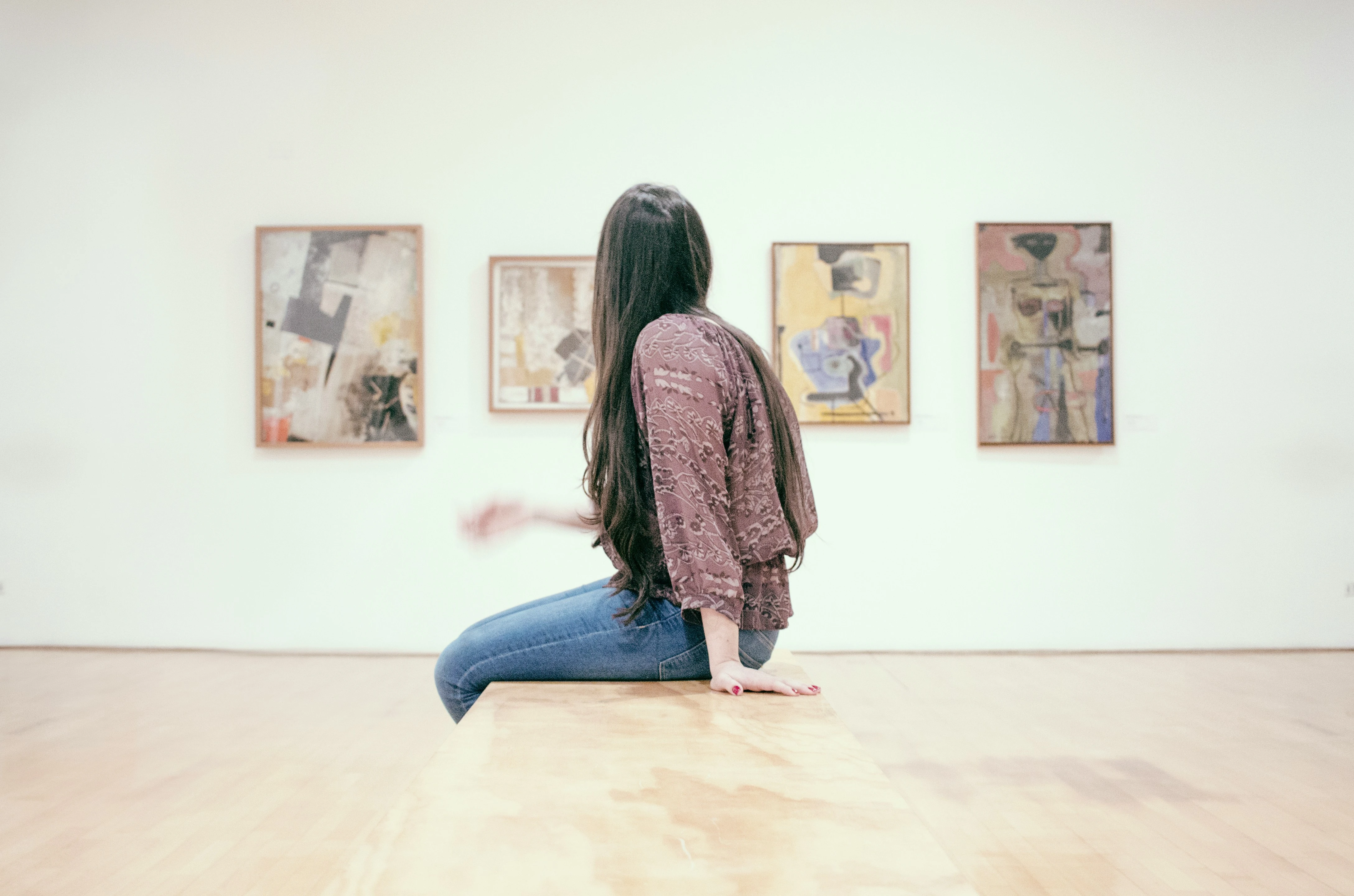 Photograph of a woman sitting on a bench in a museum gallery, looking at paintings